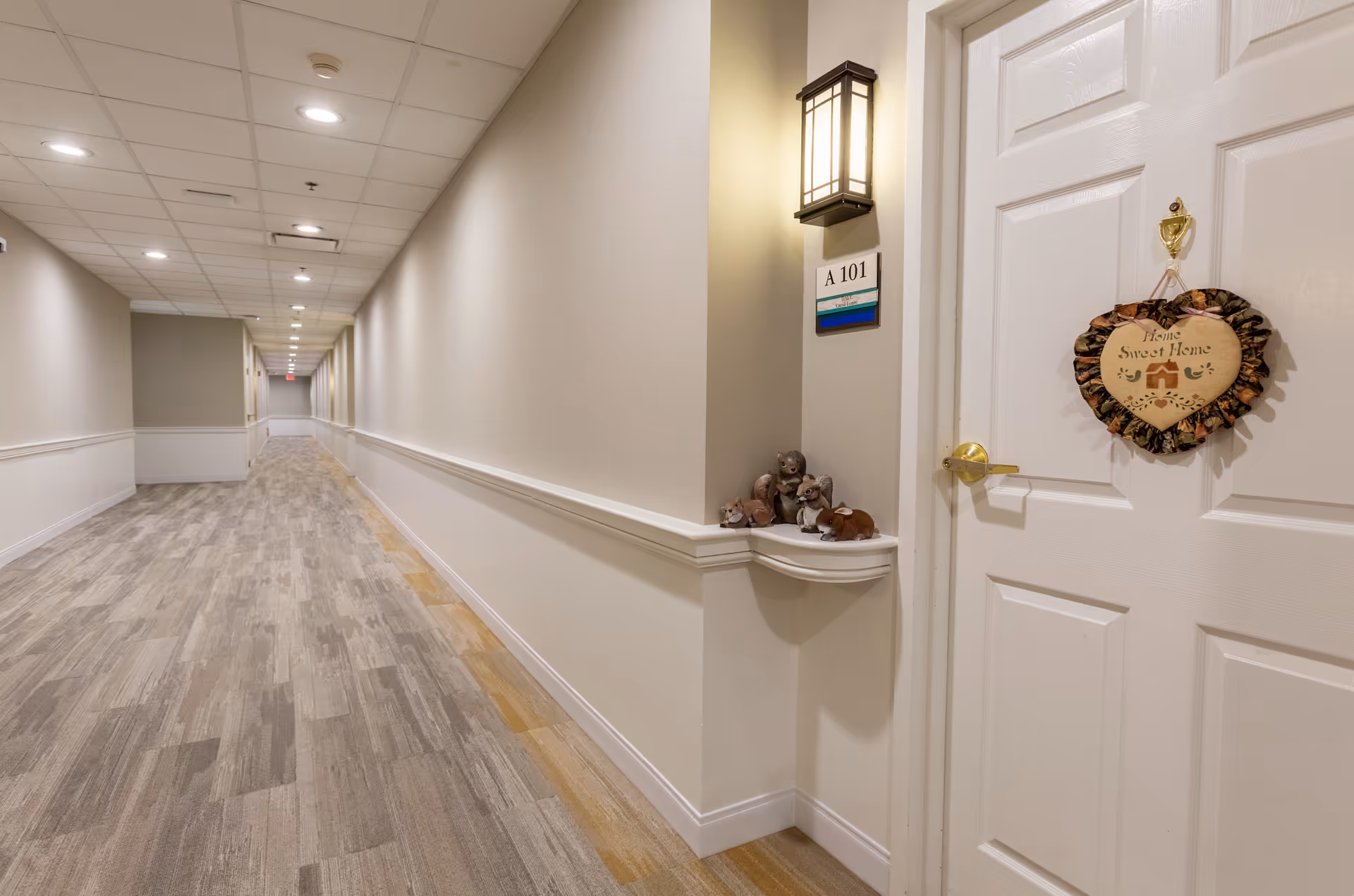 A long, well-lit hallway in a senior living facility with beige walls and wood-patterned flooring. On the right side, there is a white door with a heart-shaped wreath that reads 'Home Sweet Home'. Above the door, a wall-mounted light fixture illuminates a small shelf holding several small animal figurines. The door is labeled A 101.