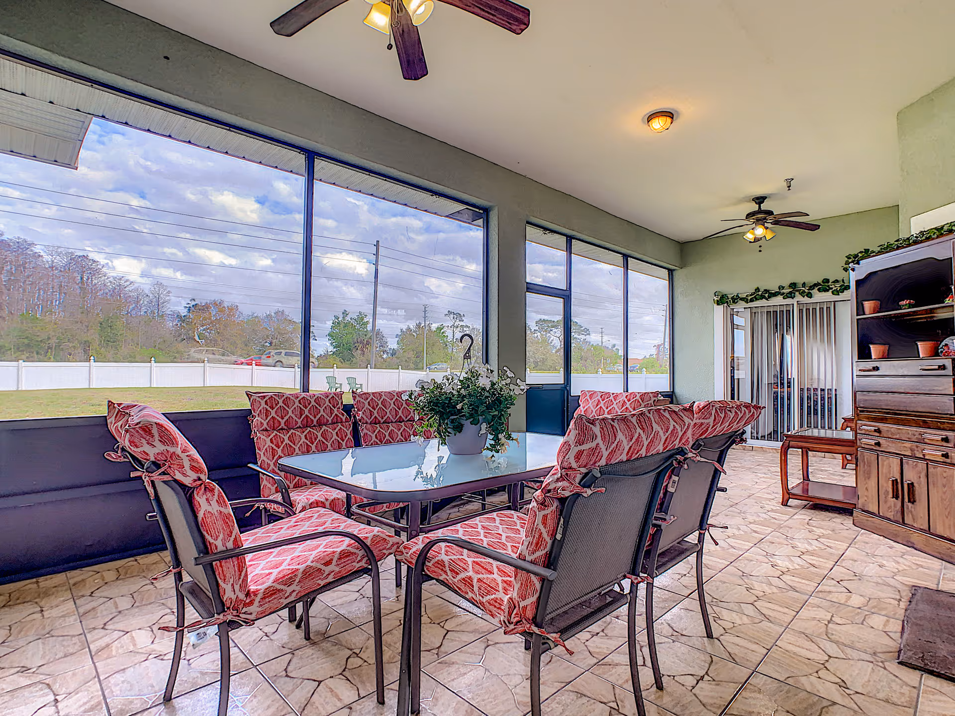 Screened sunroom with a glass-top dining table surrounded by red-patterned cushioned chairs, ceiling fans, and a view of the yard.
