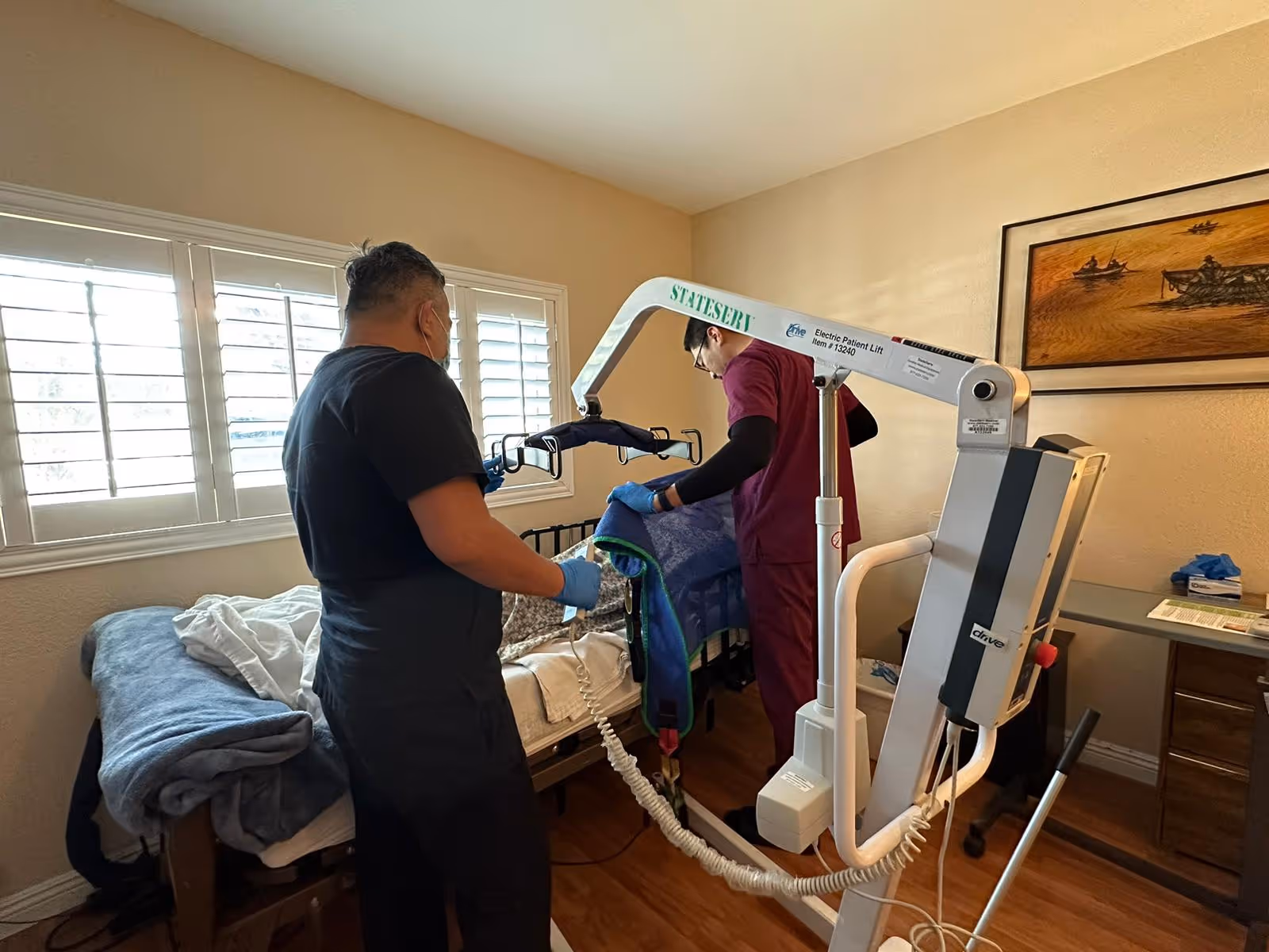 Two healthcare workers in a bedroom setting using an electric patient lift to assist a person in bed. The room has a window with white shutters, a bed with blankets, wooden flooring, and a framed painting on the wall.