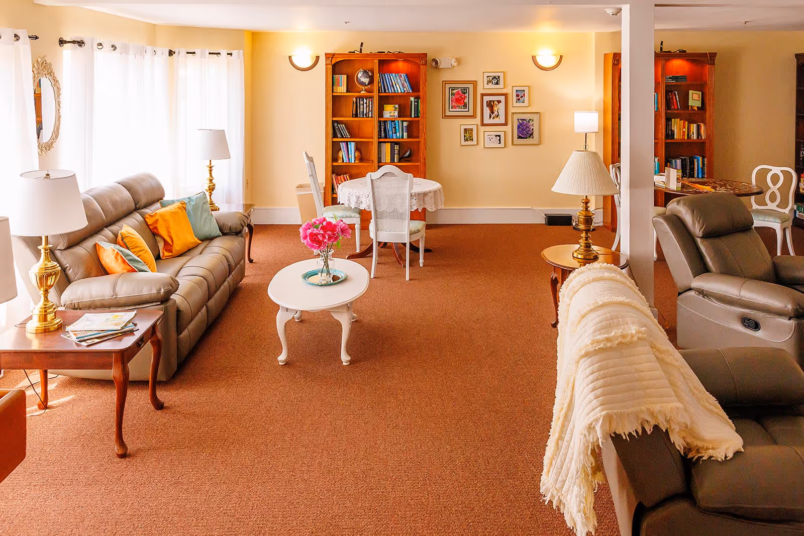 A cozy senior living room with beige walls and brown carpet. The room features a gray leather sofa with colorful pillows, a white coffee table with a vase of pink flowers, two wooden bookshelves filled with books, a small round table with a lace tablecloth and chairs, several lamps, and framed pictures on the wall.