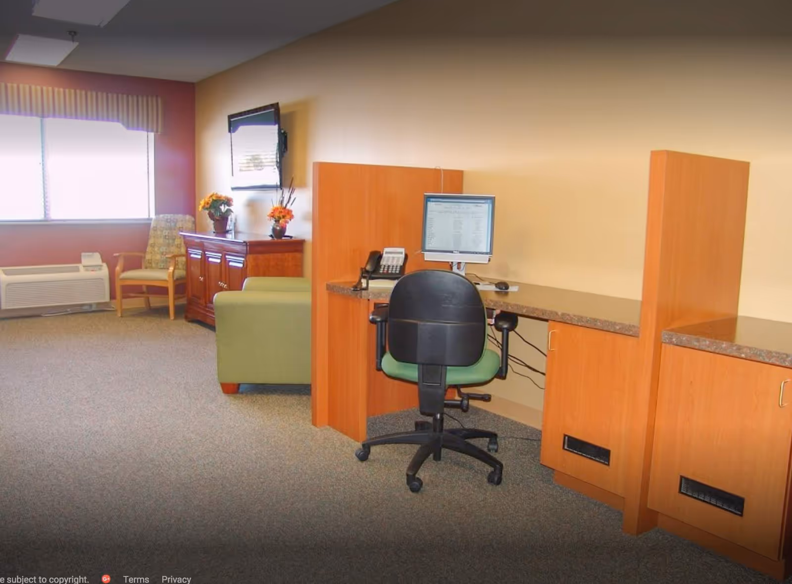 An interior room with a computer workstation featuring a black office chair and a monitor on a wooden desk with partitions. In the background, there is a green armchair, a wooden cabinet with flower vases, a wall-mounted TV, a window with striped valance, and a patterned armchair next to an air conditioning unit.