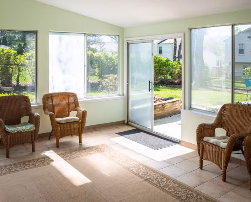 A sunlit indoor seating area with three wicker chairs, each with a cushion, placed around the edges of the room. Large windows and a sliding glass door provide a view of a green outdoor garden area with bushes and houses in the background. The floor is tiled with a decorative rug in the center.