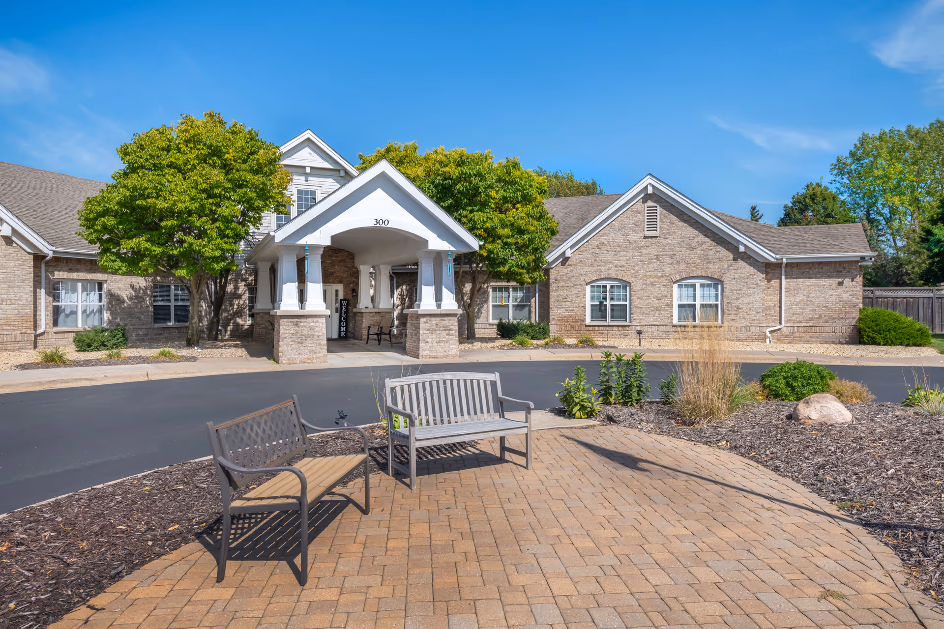 Exterior view of a senior living facility with a brick facade, a covered entrance with the number 300, two benches on a paved area in the foreground, and trees and landscaping around the building under a clear blue sky.