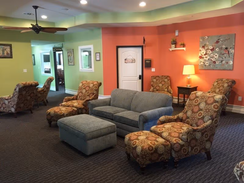 A cozy living room area in a memory care facility with a gray sofa, matching ottoman, and several patterned armchairs with ottomans. The walls are painted in green and orange tones, with a ceiling fan, a side table with a lamp, and floral artwork on the wall. A white door and an open doorway leading to another room are visible in the background.