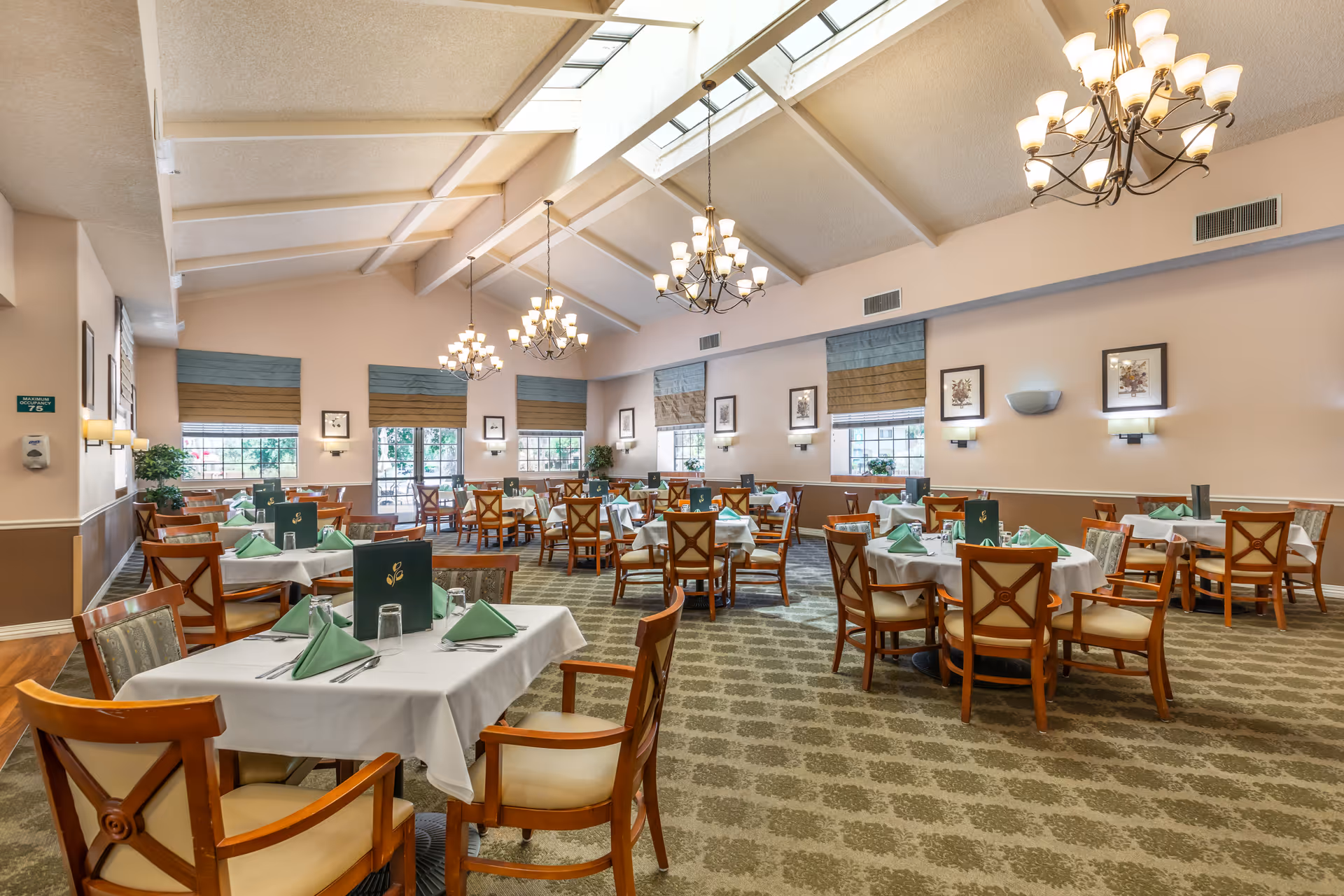 A spacious dining room with multiple tables covered in white tablecloths and set with green napkins, glassware, and menus. The room features high vaulted ceilings with skylights and multiple chandeliers. Large windows with blue and brown valances allow natural light to fill the space. The walls are decorated with framed artwork and wall sconces, and the floor is carpeted with a patterned design.