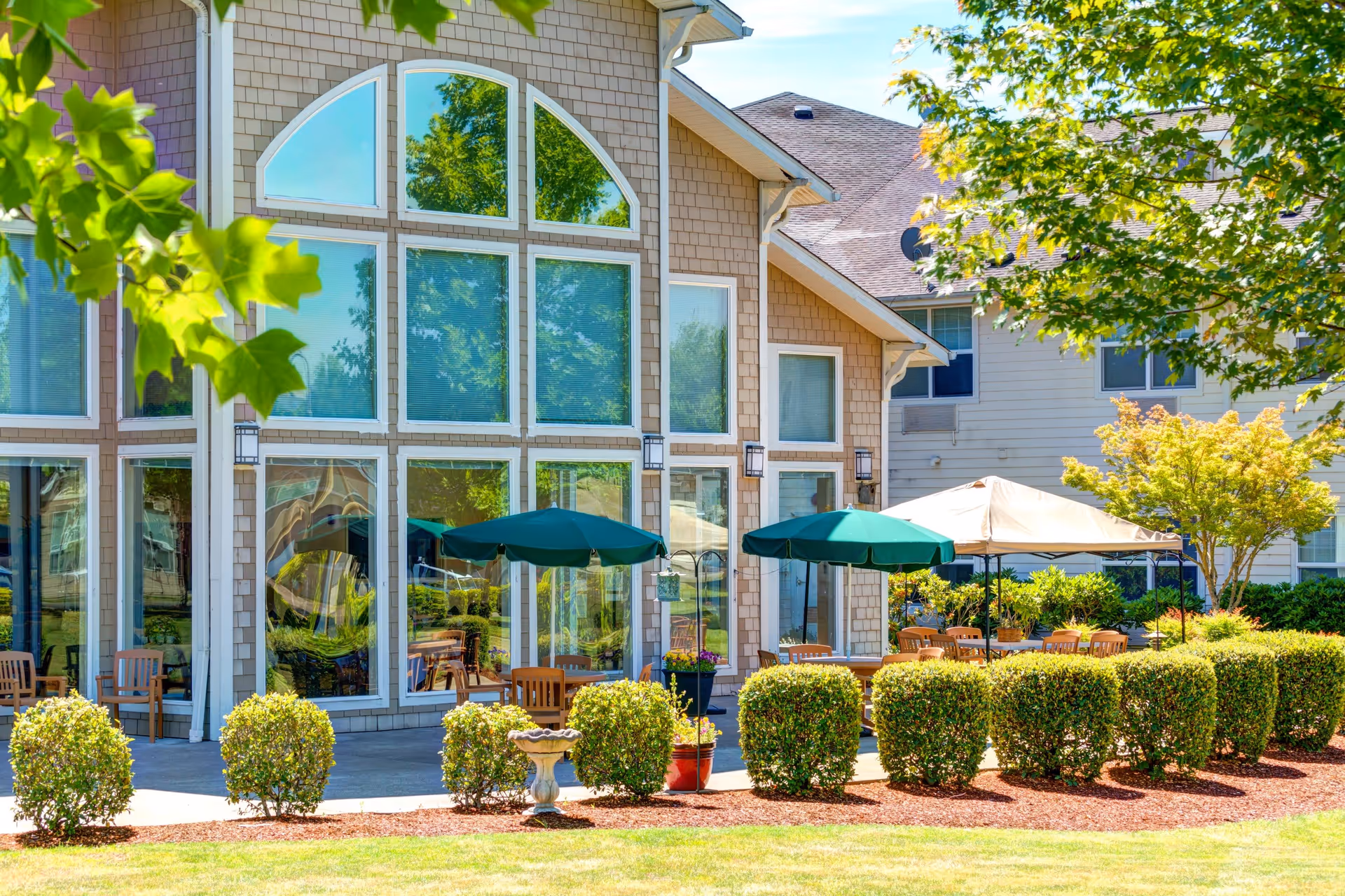 Outdoor patio area of Vineyard Heights Assisted Living with large windows reflecting trees, green umbrellas shading wooden tables and chairs, neatly trimmed bushes, and a well-maintained lawn under a sunny sky.
