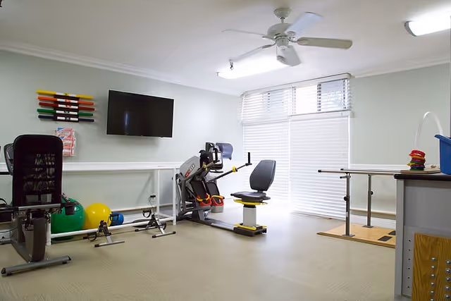 A bright exercise room with various fitness equipment including a stationary bike, resistance bands mounted on the wall, exercise balls, and parallel bars near a large window with blinds. A ceiling fan and fluorescent lights are visible on the ceiling.