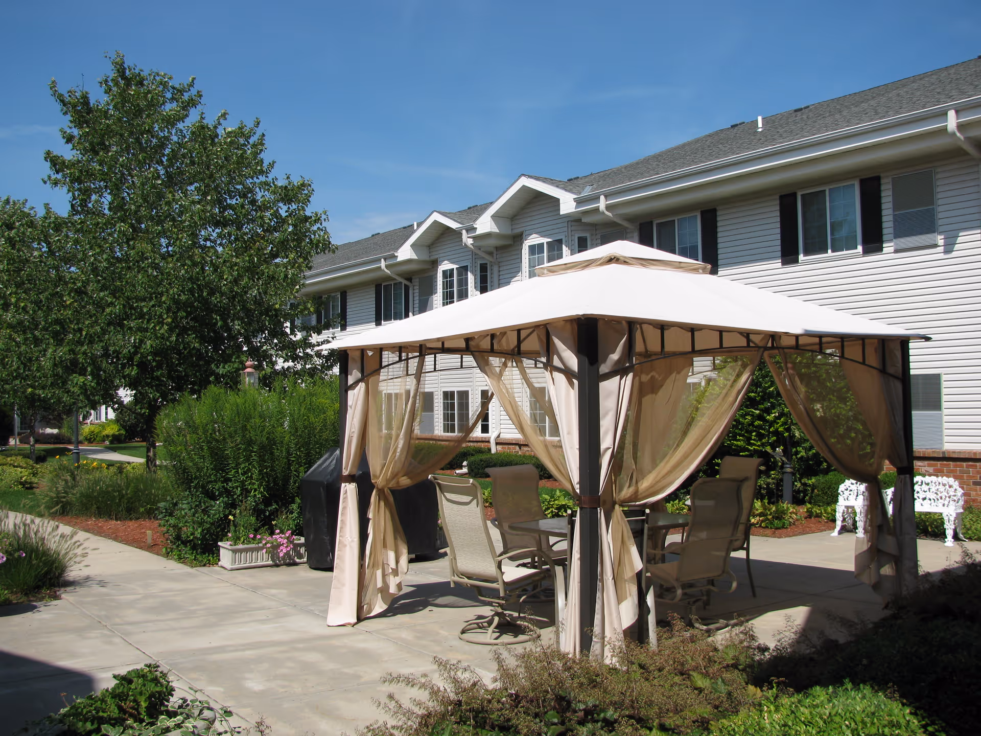 Outdoor patio area at Bailey Pointe Assisted Living at Van Dorn featuring a beige gazebo with sheer curtains, patio chairs, and a table. The area is surrounded by greenery, shrubs, and a large tree, with a multi-story building in the background under a clear blue sky.