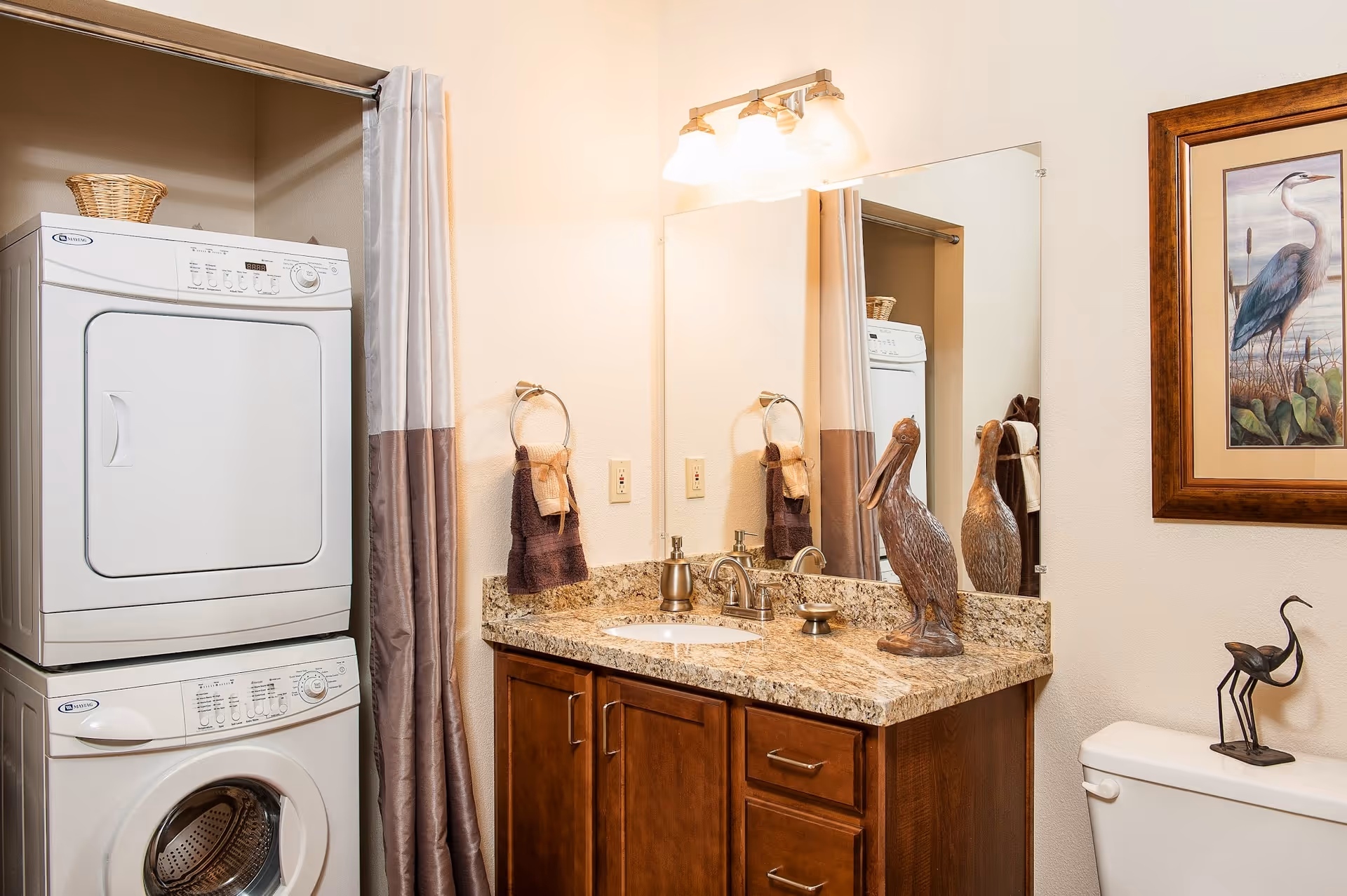 Bathroom featuring a stacked washer and dryer, a granite-top vanity with sink and mirror, toilet, and decorative artwork.