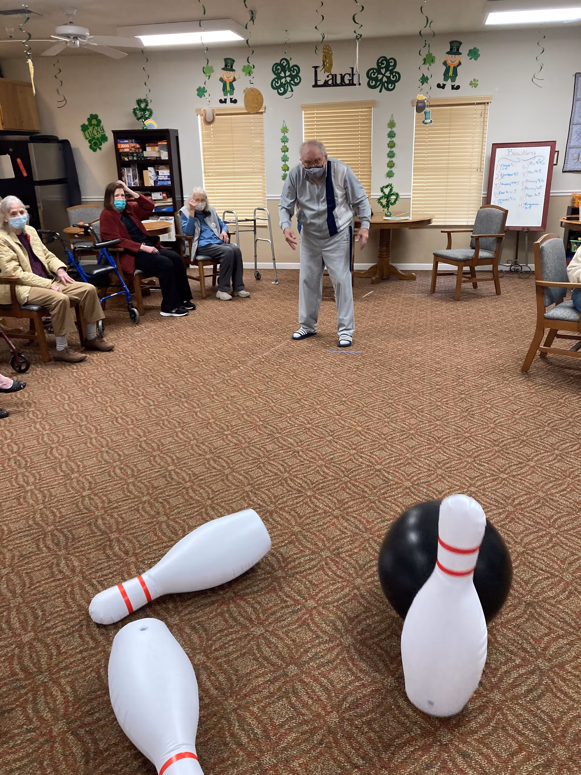 A group of elderly people sitting in chairs arranged in a semicircle in a decorated room with shamrock and leprechaun decorations. One elderly man is standing in the center, appearing to play a game of indoor bowling with inflatable pins and a black ball on the carpeted floor. Some of the seated individuals are wearing face masks.