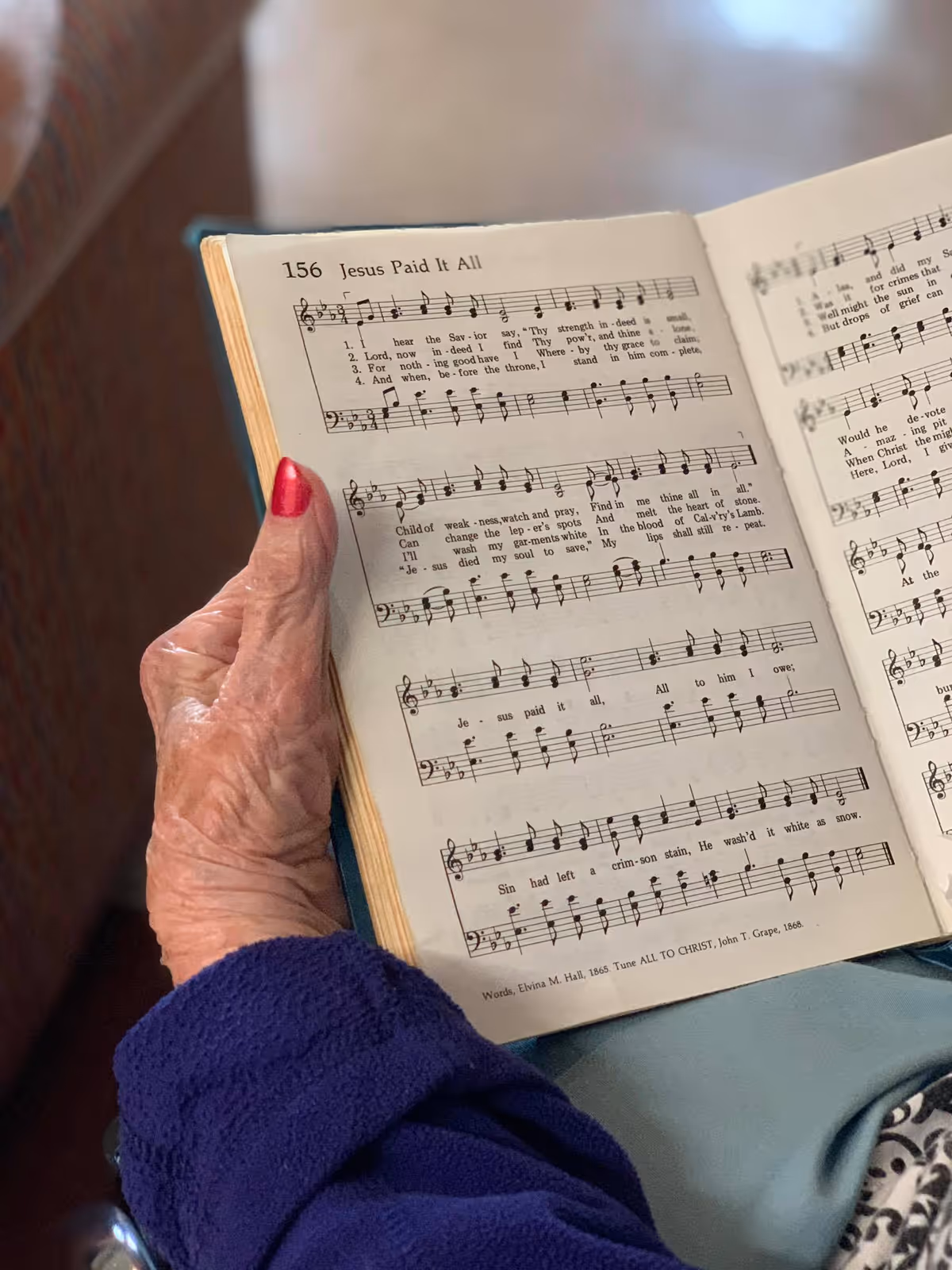 An elderly person with a purple sleeve holding an open hymnal book displaying the sheet music and lyrics for the hymn 'Jesus Paid It All'.