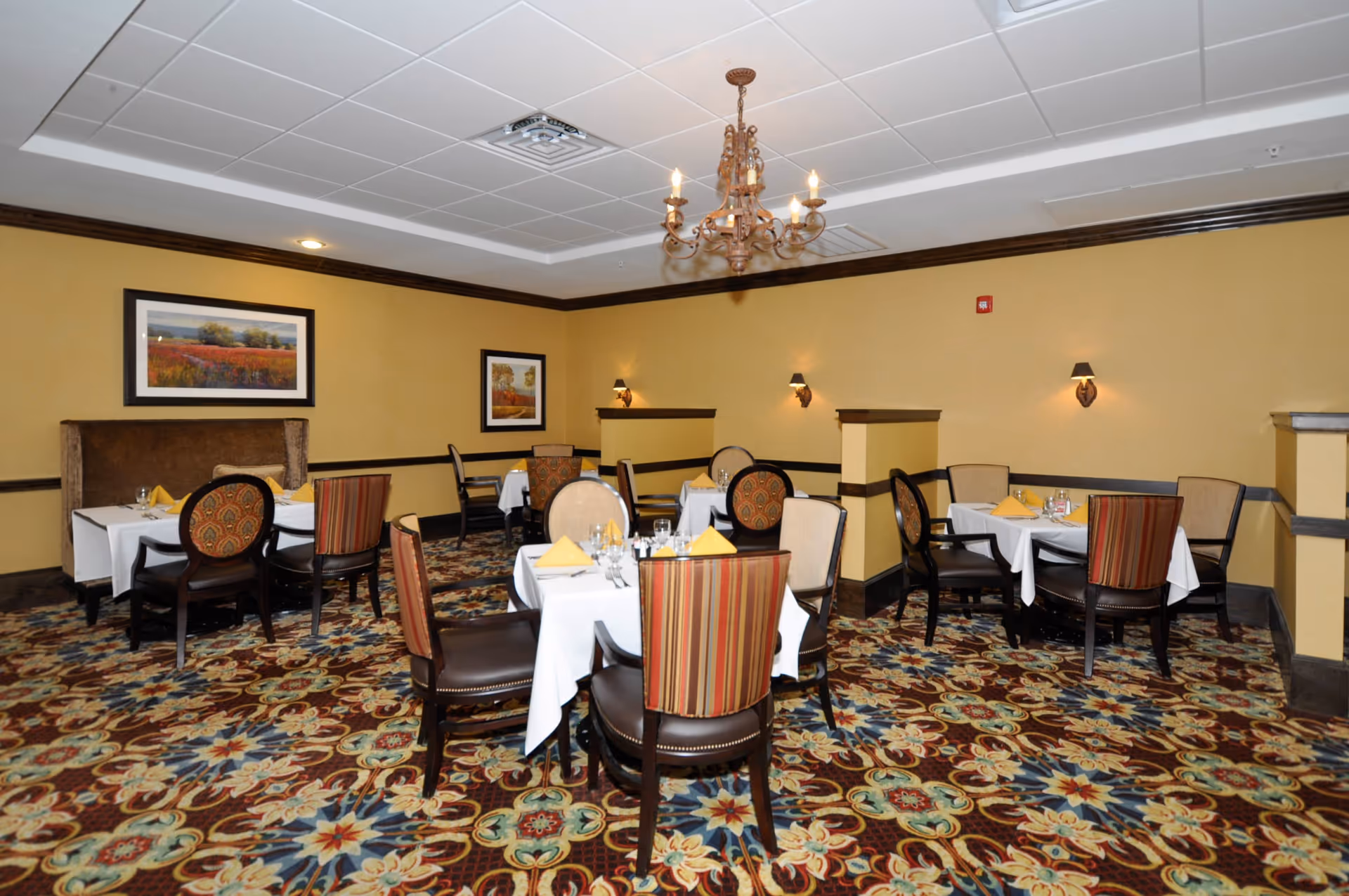 A dining room with multiple tables covered in white tablecloths, each set with yellow folded napkins, glassware, and silverware. The room has patterned carpet with floral designs, yellow walls with dark wood trim, framed landscape paintings, wall sconces, and a chandelier hanging from the ceiling.