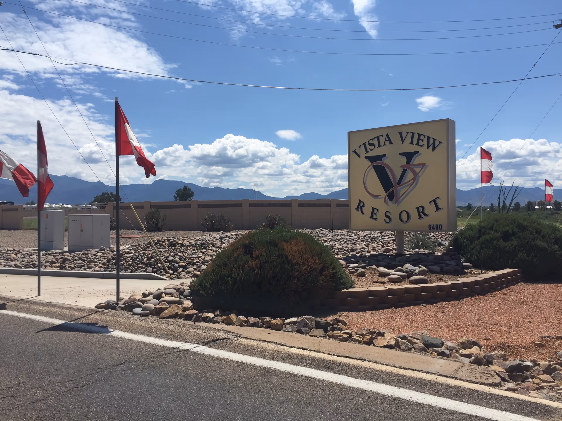 Outdoor view of the entrance sign for Vista View Resort with a landscaped area featuring rocks and bushes. Several red and white flags are displayed on poles along the roadside, with mountains and a partly cloudy sky in the background.