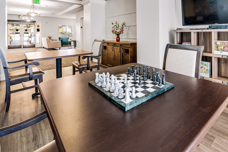 Interior view of a senior living facility common area with a dark wooden table in the foreground featuring a chessboard with chess pieces set up. Surrounding the table are chairs, and in the background, there are more tables and chairs, a wooden cabinet with a vase of flowers, a television mounted on the wall, and a seating area near the entrance with armchairs and large windows letting in natural light.