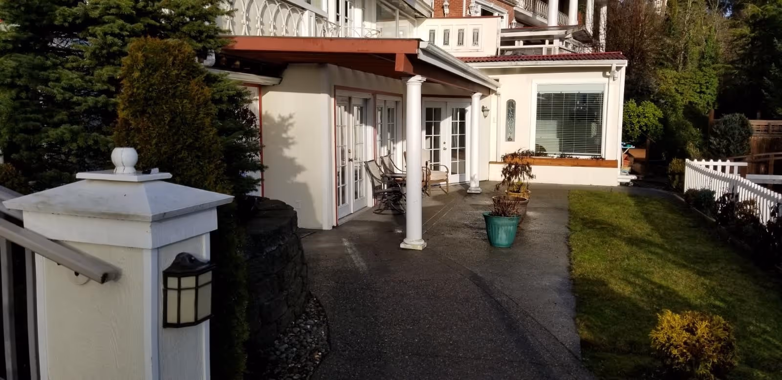 Outdoor patio area of a residential building with white columns, glass doors, potted plants, and a small lawn bordered by a white fence and shrubs.