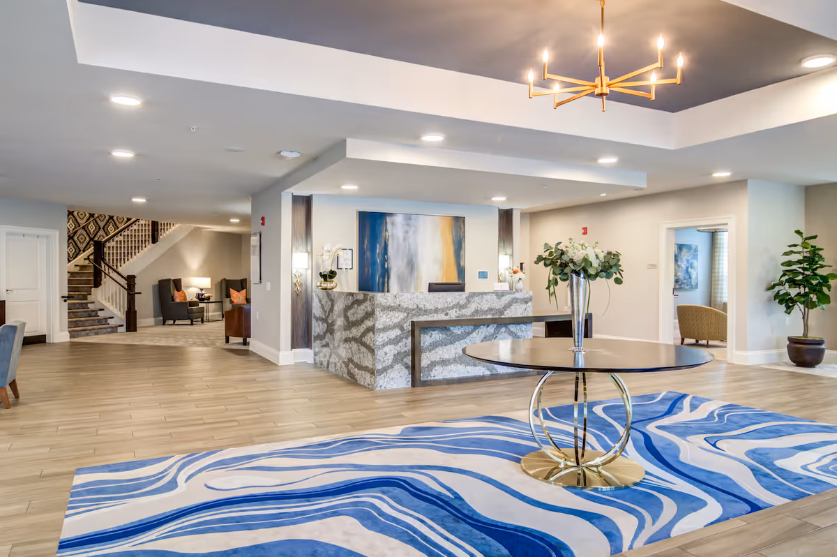 Spacious senior living lobby with a reception desk, round table with a vase on a blue patterned rug, seating areas and a staircase.