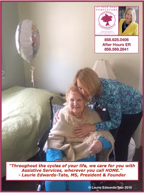 An elderly woman sitting in a wheelchair covered with a beige shawl, smiling while being embraced by a younger woman in a blue patterned top. They are in a room with a bed covered by a green blanket and a lamp on a bedside table. A silver balloon is floating near the bed.