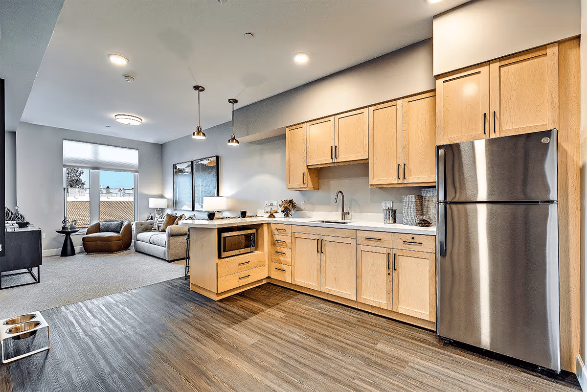 A modern open-concept living space featuring a kitchen with light wood cabinets, a stainless steel refrigerator, a built-in microwave, and a sink. The kitchen area has wood flooring that transitions to carpet in the adjacent living room, which includes a sofa, an armchair, a side table with a lamp, and large windows with blinds allowing natural light to enter.