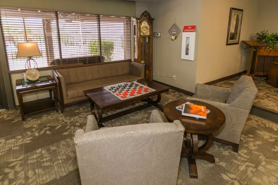 A cozy senior living common area with a brown sofa, two beige armchairs, a wooden coffee table with a checkers game set, and a side table with a lamp and decorative items. A grandfather clock stands against the wall near a fire extinguisher cabinet, and a framed picture hangs on the wall. The room has large windows with blinds and patterned carpet flooring.