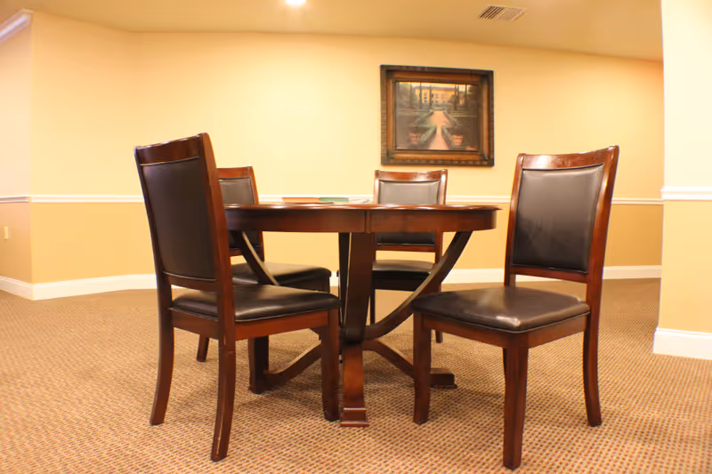 A small round wooden table with four dark wooden chairs with black cushioned seats and backs, set in a carpeted room with beige walls. A framed painting hangs on the wall behind the table.