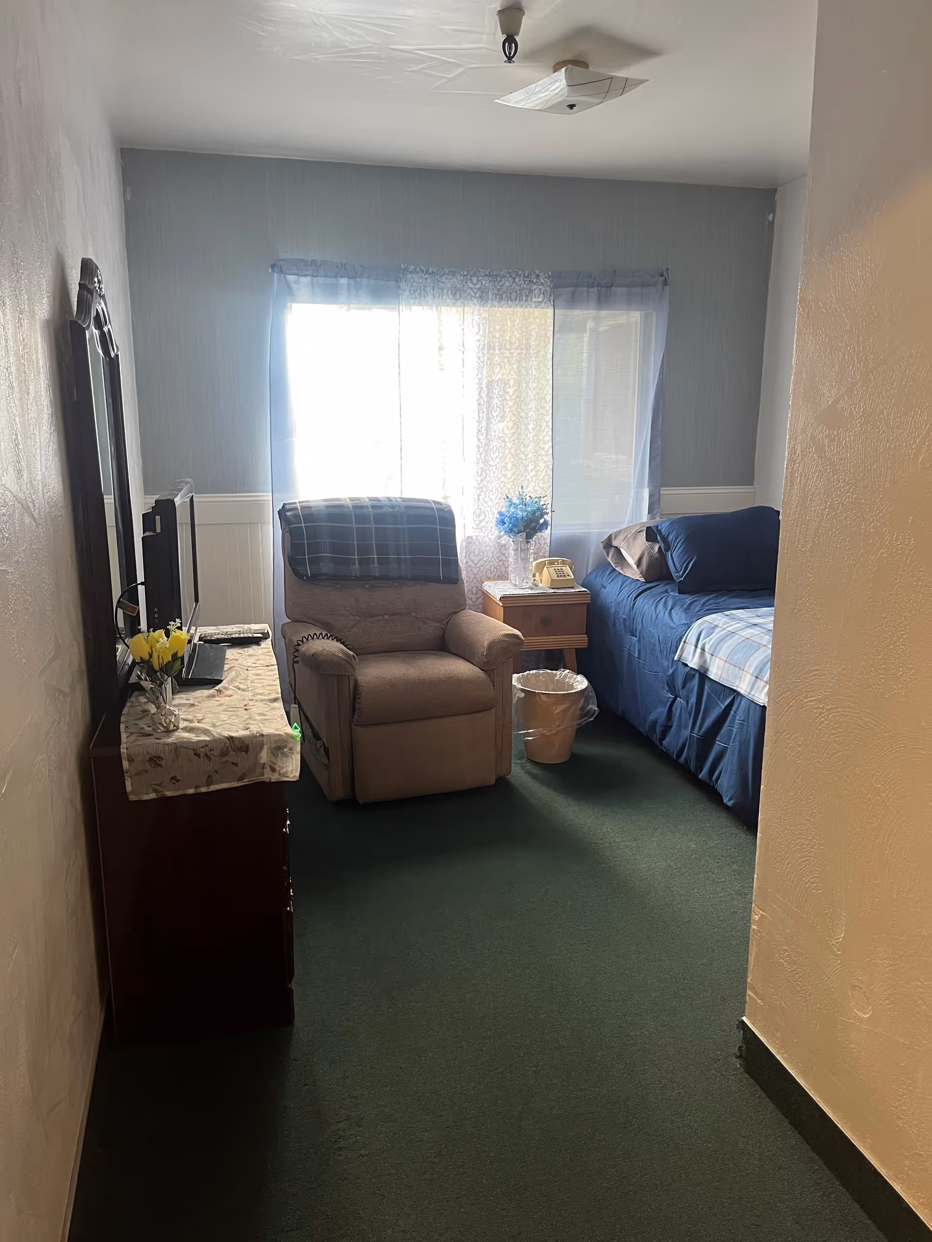A small, cozy bedroom with a single bed covered in blue bedding, a beige recliner chair with a plaid blanket draped over the back, a wooden nightstand with a yellow rotary phone and a vase of blue flowers, a dresser with a floral cloth and a small TV on top, and a window with sheer curtains letting in natural light.