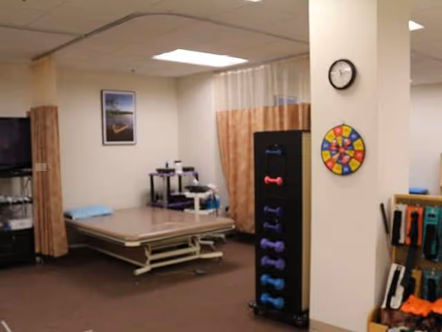 Rehabilitation/therapy room with a bed, weight rack, curtains, and activity wall at Laurelhurst Village.