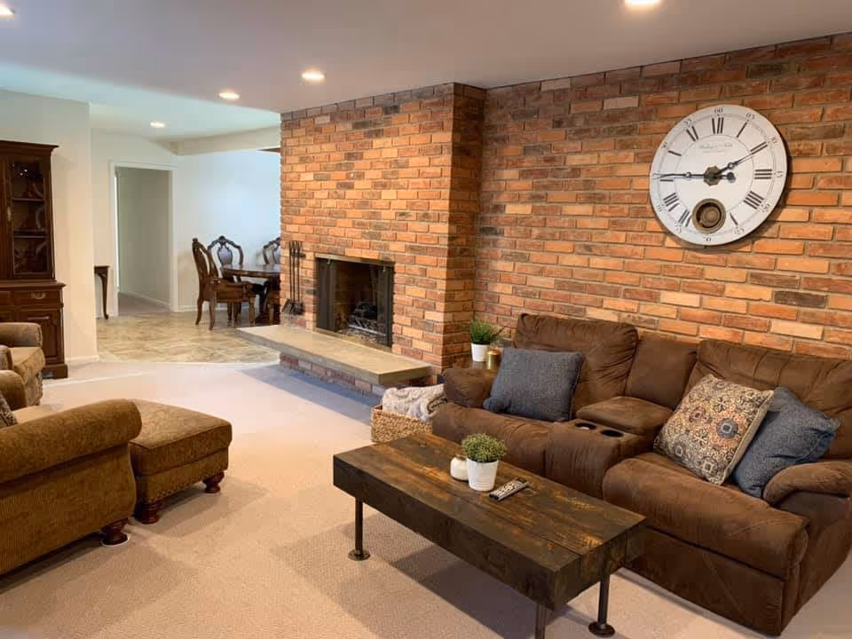 Cozy living room with a brown sofa, wooden coffee table, brick fireplace wall topped by a large round clock, and a dining area visible in the background.
