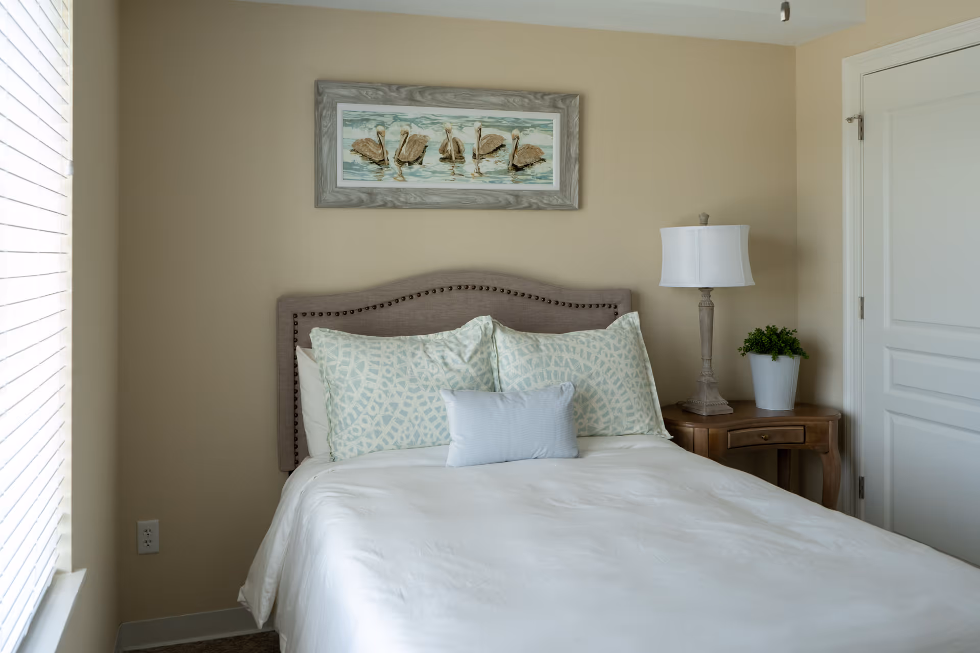 A neatly made bed with a beige upholstered headboard, white bedding, and three pillows in a bedroom. A wooden nightstand with a white lamp and a small green plant is next to the bed. Above the bed hangs a framed picture of pelicans on water. A window with blinds is on the left side, and a closed white door is on the right.
