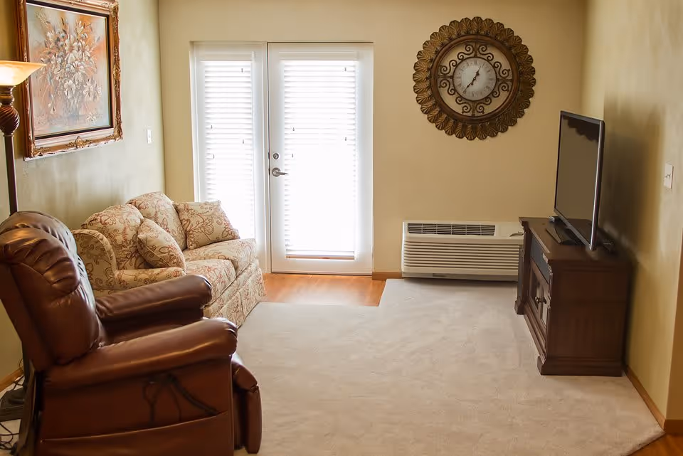 A cozy living room with a brown leather recliner chair and a floral patterned sofa with cushions. There is a wooden TV stand with a flat-screen TV on it, a large decorative wall clock, a floor lamp, and a framed painting on the wall. The room has beige walls, a light carpet, and a door with white blinds letting in natural light.