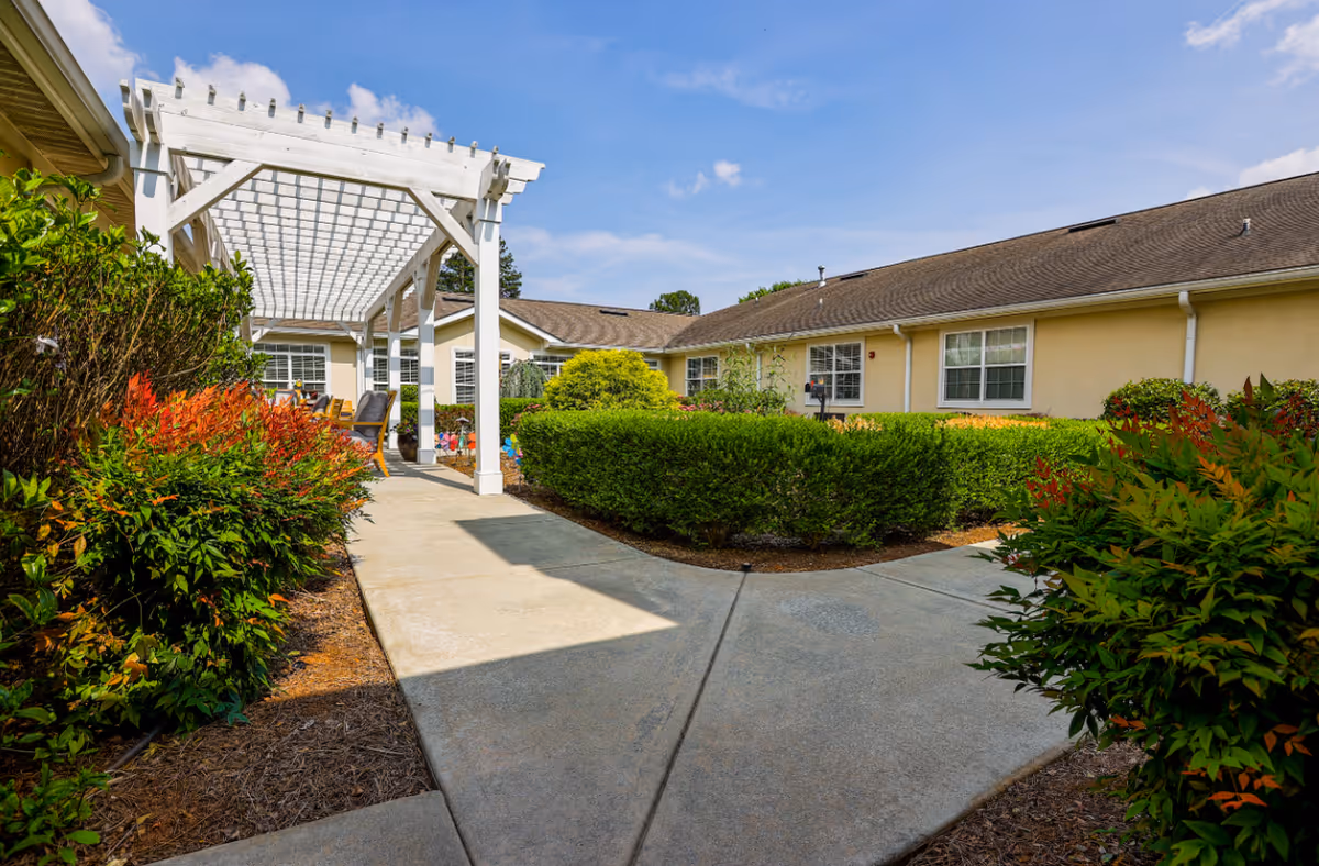 Outdoor walkway at Addington Place of Johns Creek with a white pergola, surrounded by green bushes and colorful plants, leading to a beige building under a blue sky with some clouds.