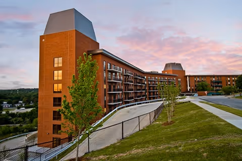Exterior view of a multi-story senior living facility building with brick walls and balconies, surrounded by green grass and trees under a partly cloudy sky during sunset.