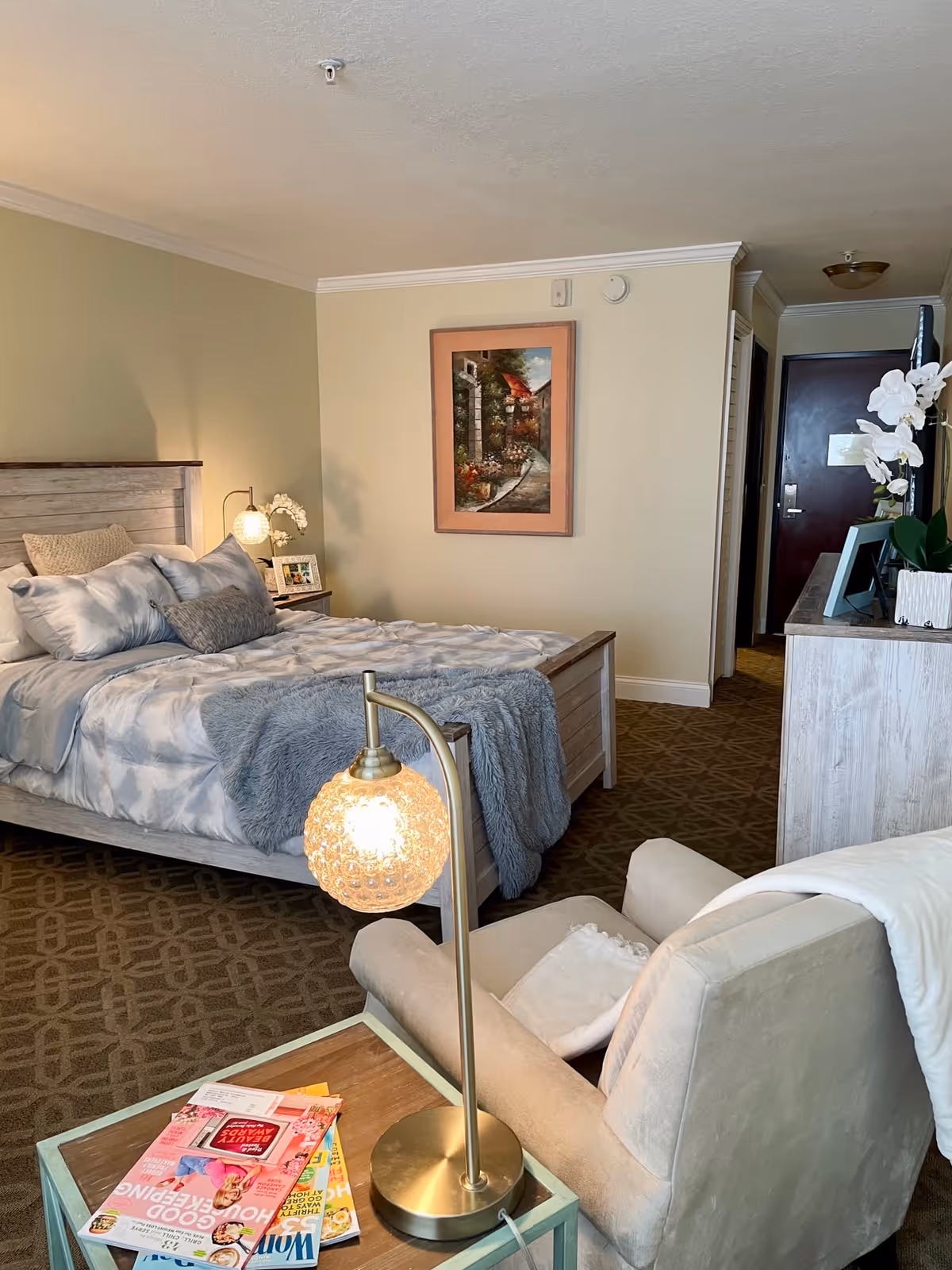 A cozy bedroom in Grace Manor Suites featuring a neatly made bed with gray and white bedding, a soft gray throw blanket, and multiple pillows. Next to the bed is a small nightstand with a lamp and a framed photo. In the foreground, there is a beige armchair with a white throw draped over it, positioned next to a small table holding a brass lamp with a textured glass globe and several magazines. The room has beige walls, a framed painting, patterned carpet, and a dresser with a white orchid and picture frames. The entrance door is visible in the background.