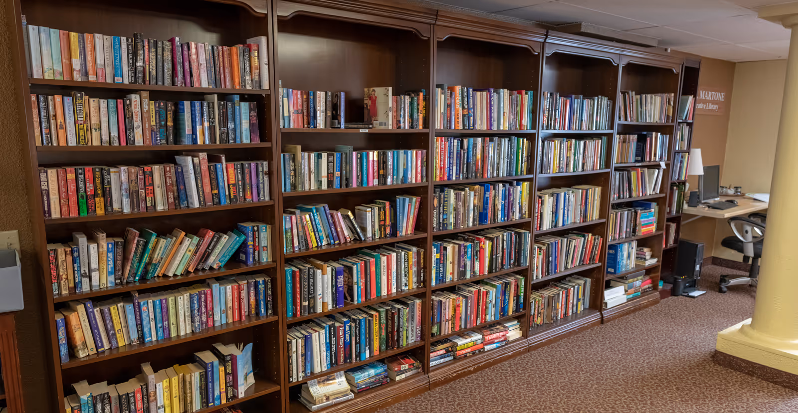 Interior view of a library room with multiple wooden bookshelves filled with a variety of books. There is a computer workstation with a chair and a lamp in the background. The carpet is patterned and the walls are painted in warm tones. A sign on the wall reads 'MARTONE Library'.
