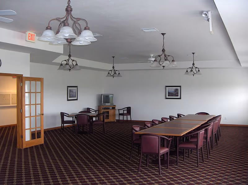 A large room with a patterned carpet, several rectangular tables arranged in a row with purple chairs around them, and additional smaller tables with chairs near the back wall. The walls are white with two framed pictures, and there is a small wooden cabinet with a television on top. Multiple ceiling light fixtures and ceiling fans are visible, and a wooden door with glass panels is open on the left side.