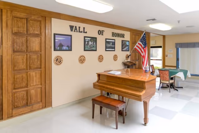 Interior view of a senior living facility featuring a wooden piano with a bench, an American flag on a stand, and a 'Wall of Honor' display with framed photos and circular plaques on a beige wall. There is a wooden door on the left and a small table with chairs near a window with vertical blinds on the right.