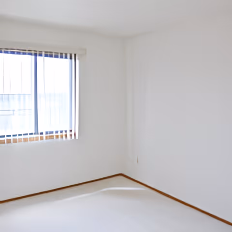 Empty white bedroom with a window fitted with vertical blinds and light wood baseboards.
