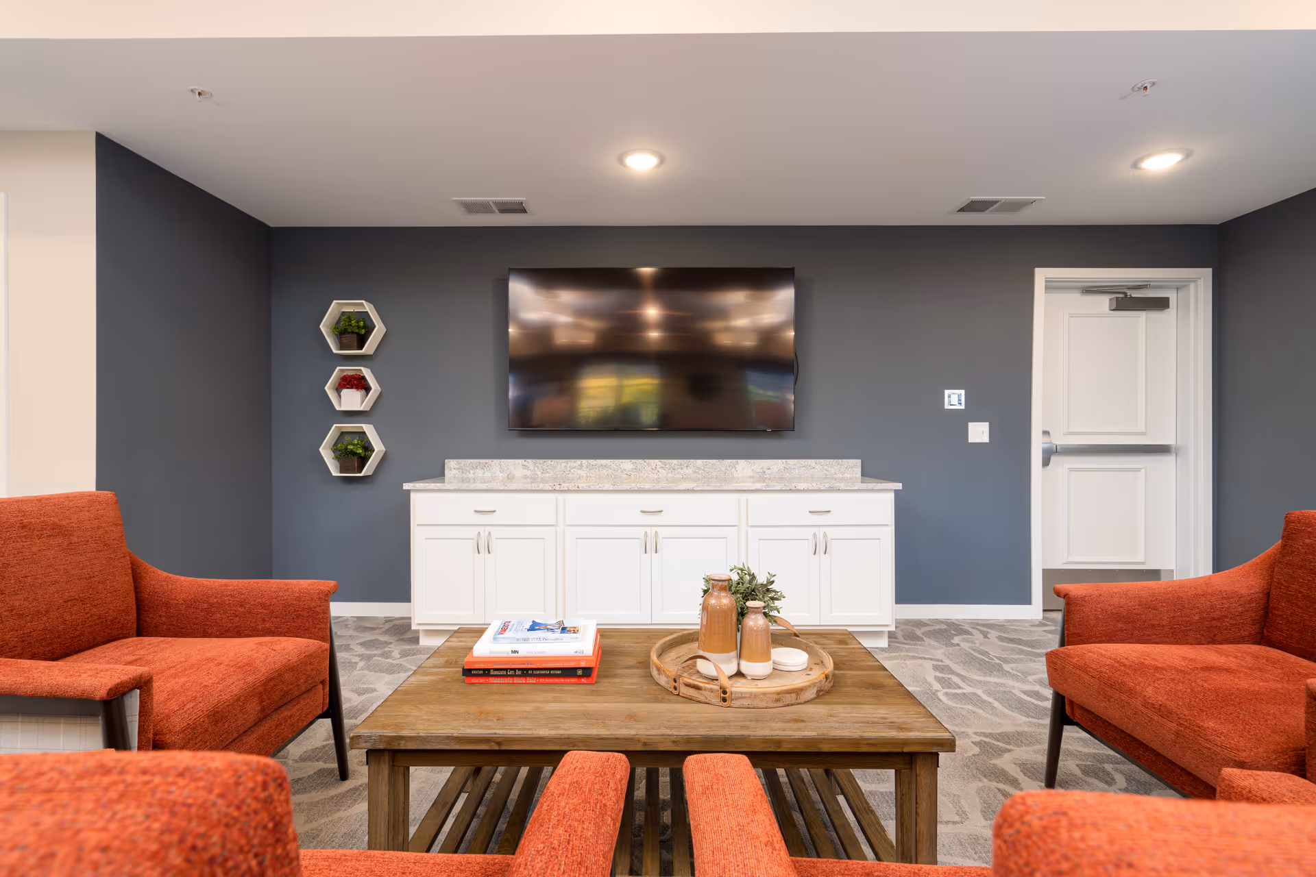 A cozy living room area with four orange upholstered chairs arranged around a wooden coffee table. On the table are decorative items including books and vases on a tray. Against a dark gray wall is a white cabinet with a mounted flat-screen TV above it. Three small hexagonal shelves with plants are mounted on the left side of the wall. A white door is visible on the right side of the room.