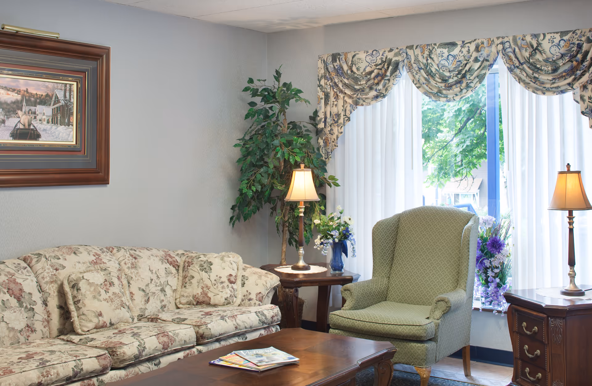 A cozy living room area with a floral patterned sofa, a green upholstered armchair, two wooden side tables each with a lamp, a coffee table with magazines, a large window with sheer white curtains and floral valances, a potted plant, and a framed painting on the wall.
