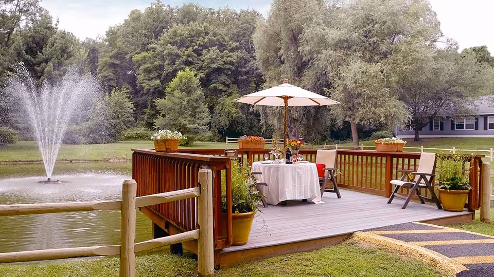 A wooden deck overlooking a pond with a water fountain, featuring a table covered with a white tablecloth, an umbrella, chairs, and potted plants, surrounded by green trees and grass.