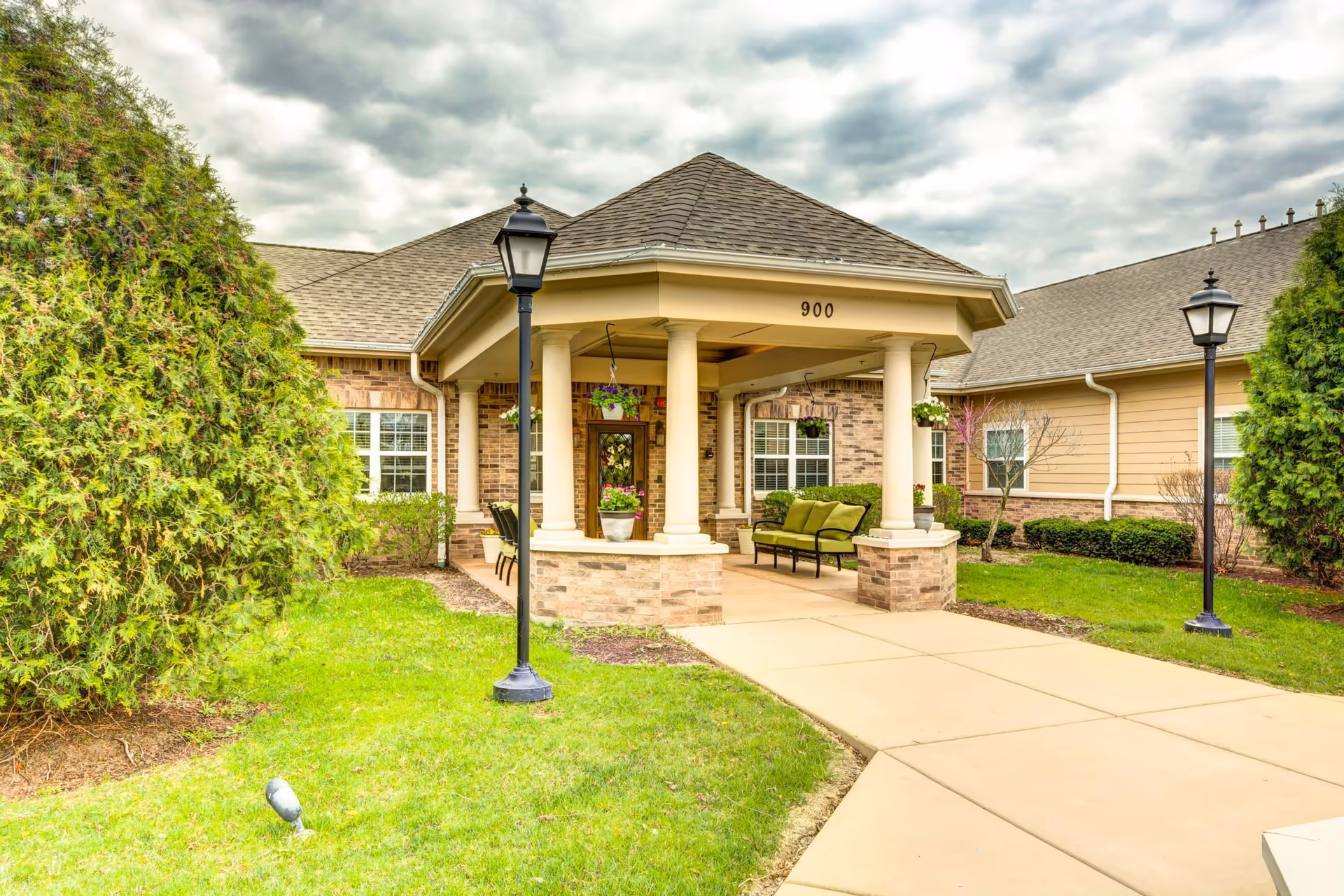 Covered entrance with columns and seating leading to the front door of a single-story brick building, flanked by lamps and landscaping.