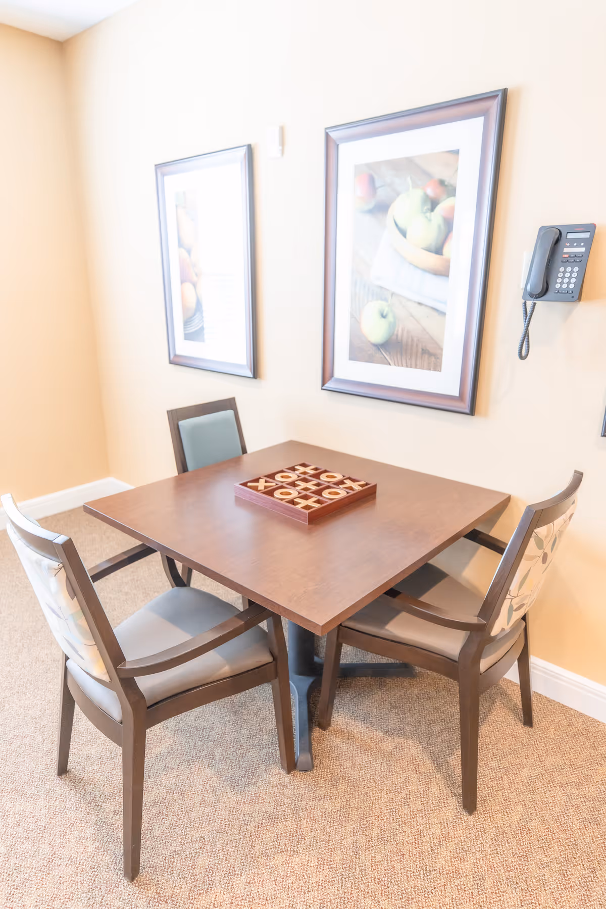 A small square wooden table with a tic-tac-toe game set on top, surrounded by four chairs with cushioned seats and backs. The room has beige walls with two framed pictures of fruit hanging on the wall, and a wall-mounted telephone is visible on the right side.