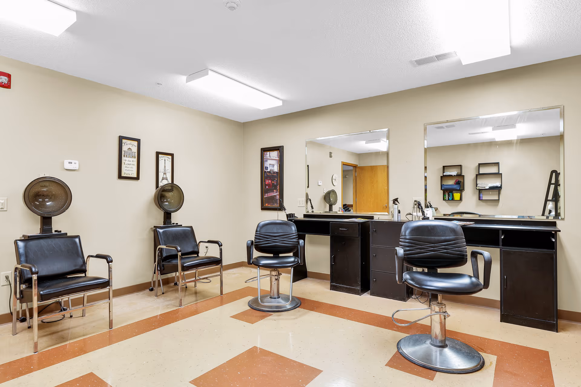 Interior of a hair salon area in an assisted living facility featuring two black salon chairs in front of large mirrors and two black chairs with vintage hair dryers along the wall. The room has beige walls, framed pictures, and a tiled floor with a brown and beige pattern.