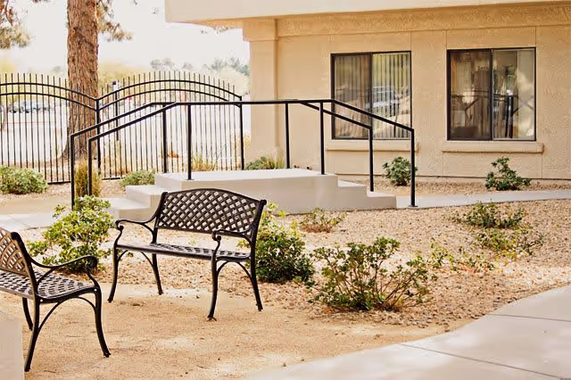 Courtyard outside a facility with metal benches, gravel landscaping, a wheelchair ramp with handrails, and building windows.