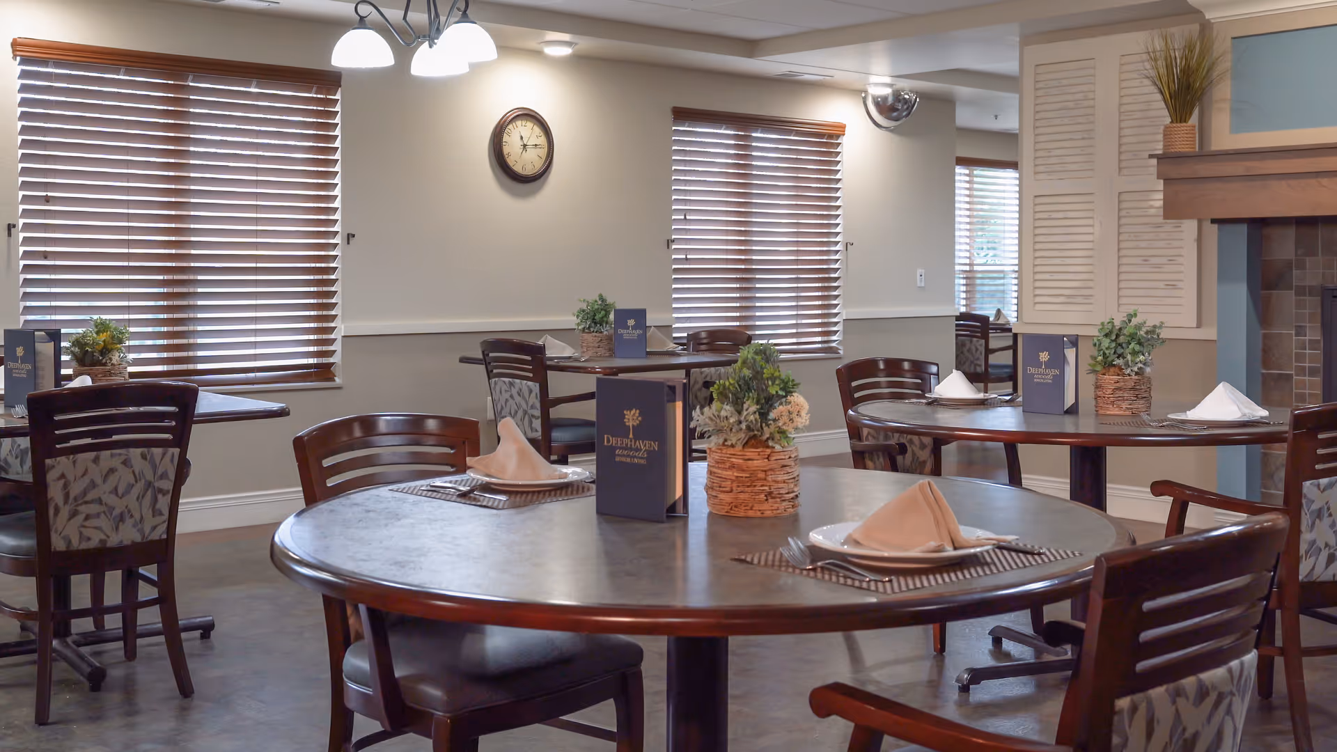 A dining room in Deephaven Woods Senior Living with round wooden tables set with plates, napkins, and silverware. Each table has a small floral centerpiece and a menu. The room has windows with wooden blinds, a wall clock, and soft lighting from ceiling fixtures.