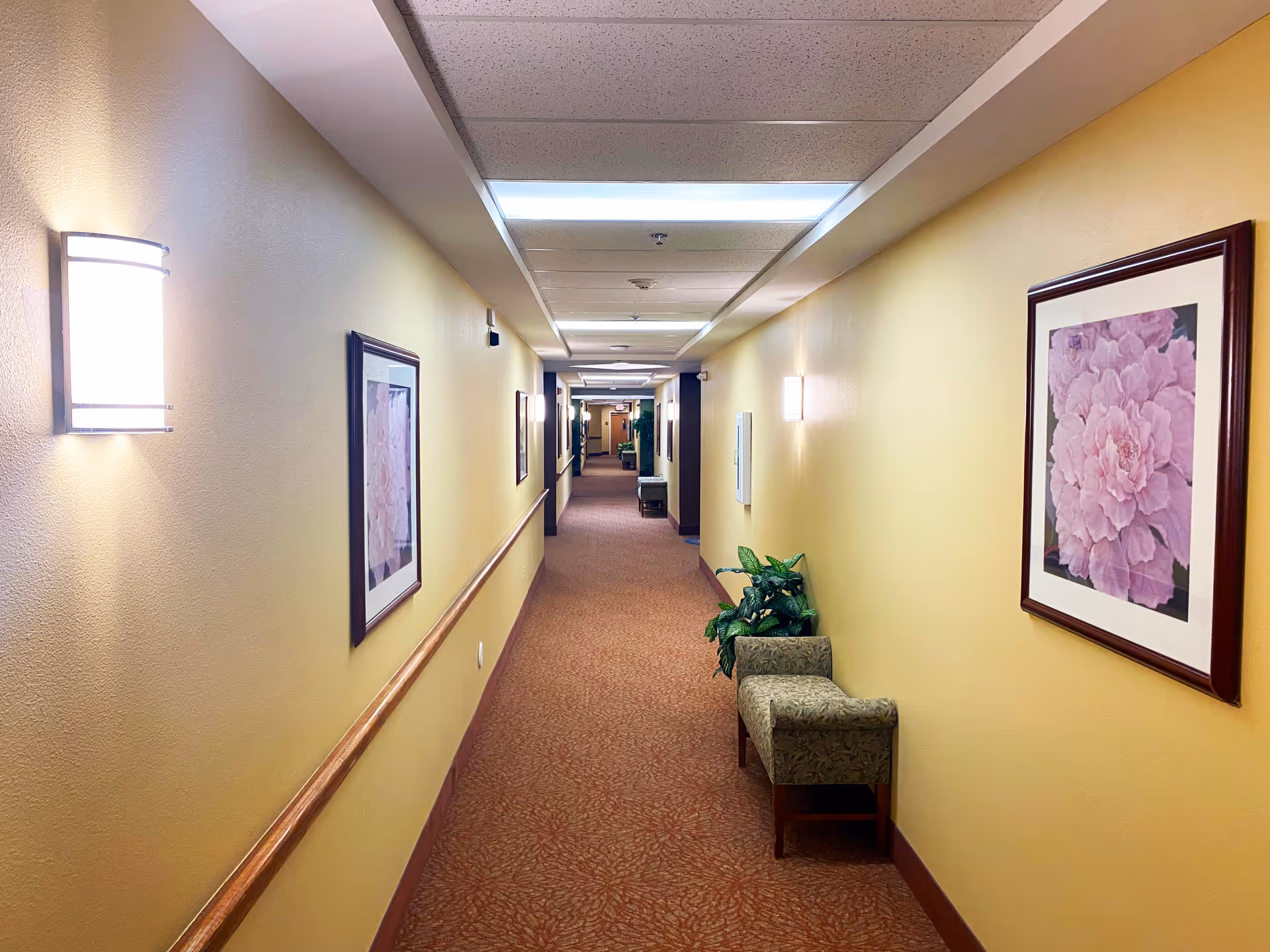A long, well-lit hallway in a senior living facility with framed artwork and seating.