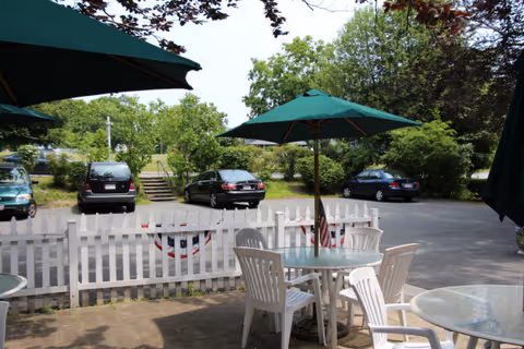 Outdoor seating area with white plastic chairs and glass tables under green umbrellas, a white picket fence decorated with patriotic bunting, and a parking lot with several cars and green trees in the background.