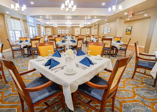 An elegant dining room with multiple tables set with white tablecloths, blue napkins, and wooden chairs under chandeliers.