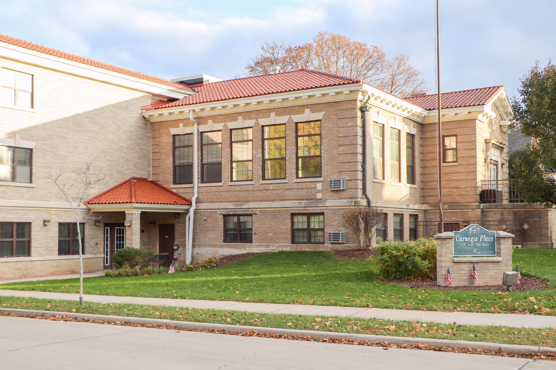 Exterior view of Carnegie Place, a brick building with large windows and a red-tiled roof. There is a green lawn with a sidewalk in front and a sign that reads 'Carnegie Place 108 South 20th Street' with small American flags around it.