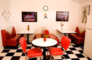 A retro-style dining area with red cushioned booths and chairs around white tables. The floor has a black and white checkered pattern. There are framed pictures and a clock on the walls, along with small vases holding flowers on the tables.
