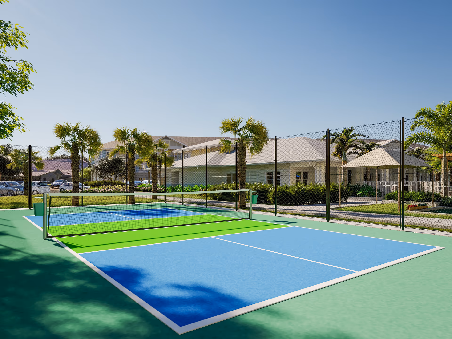Outdoor tennis court with a net, surrounded by a chain-link fence and palm trees. In the background, there are buildings with light-colored roofs and a clear blue sky.
